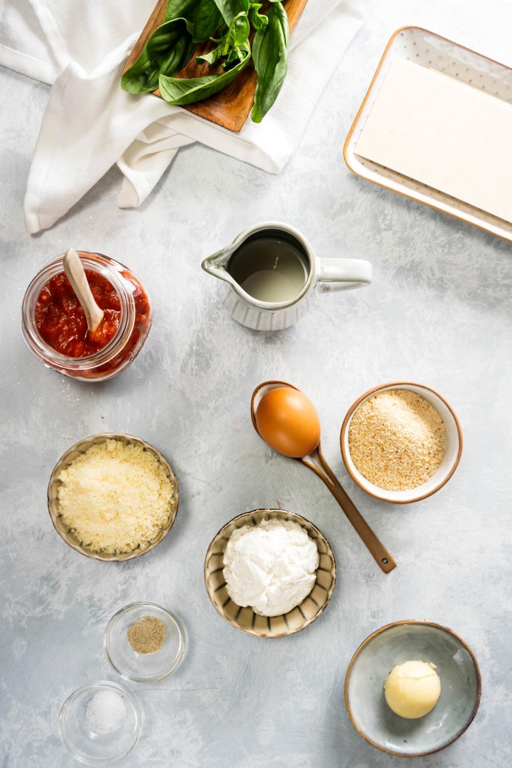 A bowl with a mozzarella cheese ball in it, a bowl of ricotta cheese, a bowl of Parmesan cheese, a bowl of breadcrumbs, an egg, a jar of tomato sauce, and a picture of water all on a gray counter.