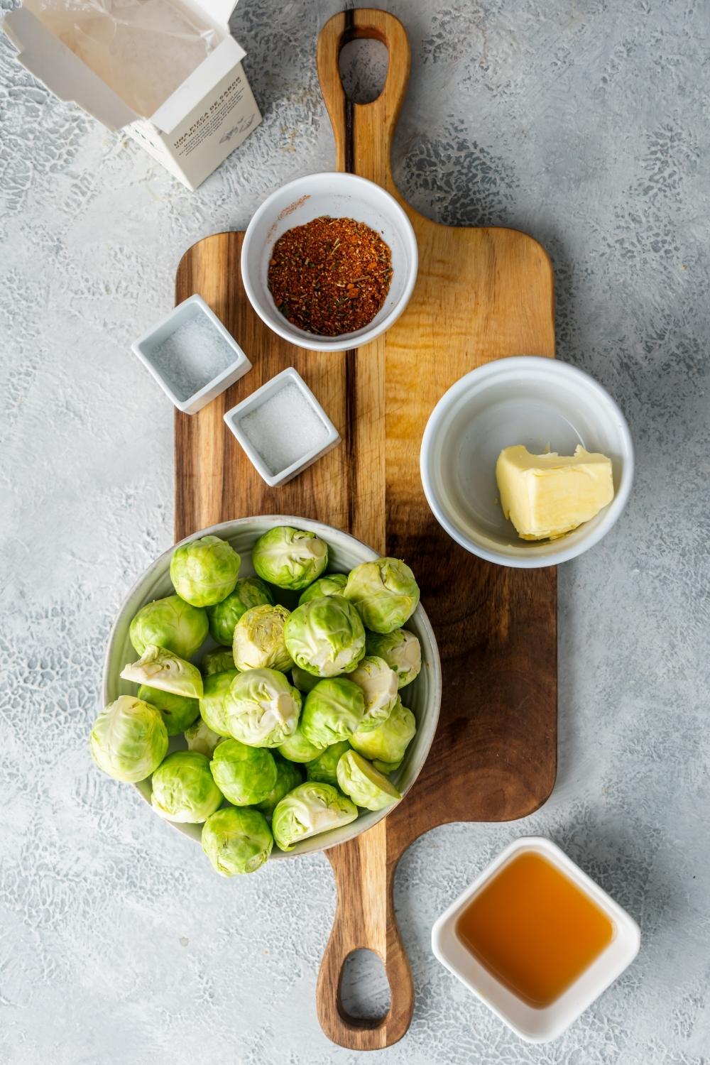 A bowl of brussel sprouts, butter in a bowl, and seasoning in a bowl on a wooden cutting board. In front of it is honey in a bowl.