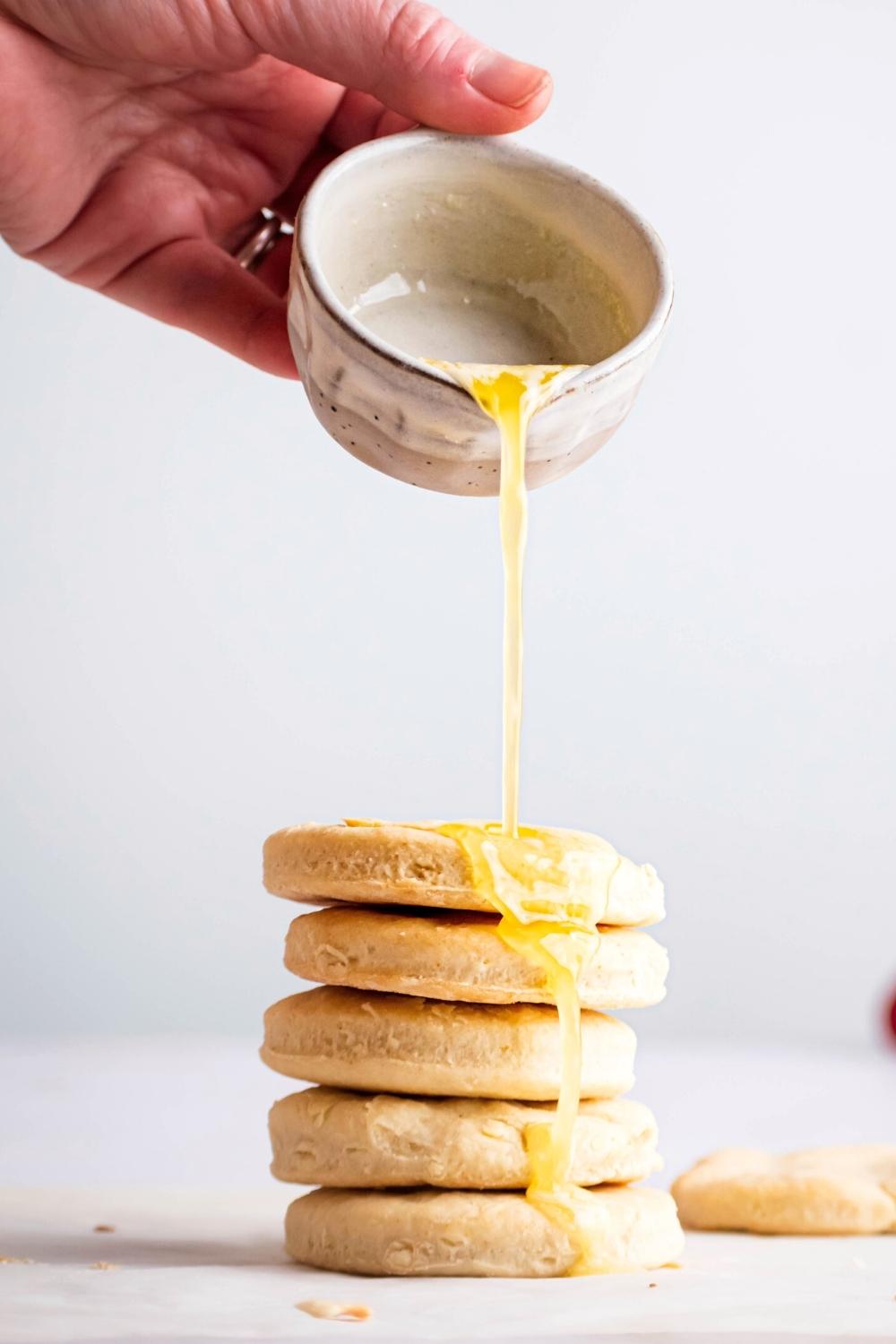 A hand pouring melted butter on top of five biscuit stacked on top of one another.