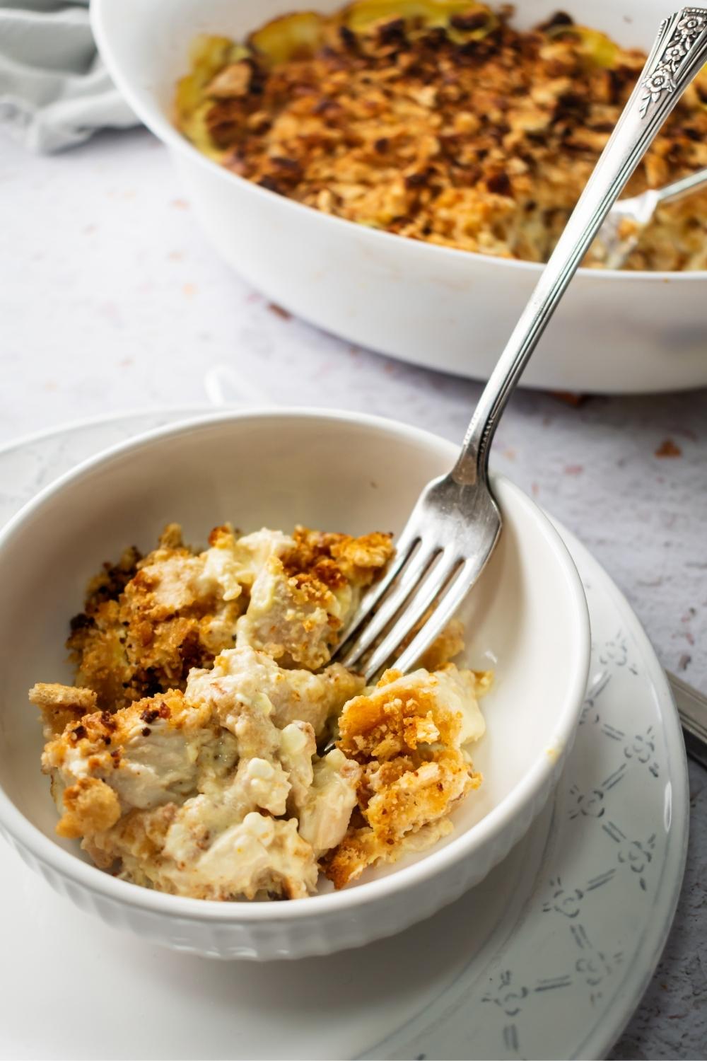 Cheesy chicken pieces with crushed up Ritz crackers on it in a white bowl that has a fork in it. Behind it is part of a casserole dish filled with million dollar casserole.