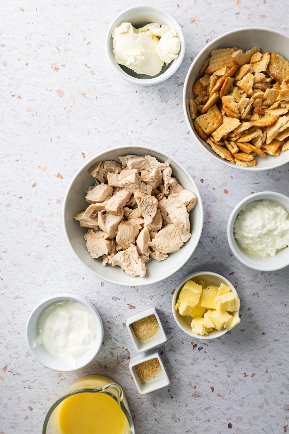 A bowl of sour cream, a bowl of butter, a bowl of cottage cheese, a bowl of chicken pieces, a bowl of crushed Ritz crackers, and a bowl of cream cheese all on a white counter.
