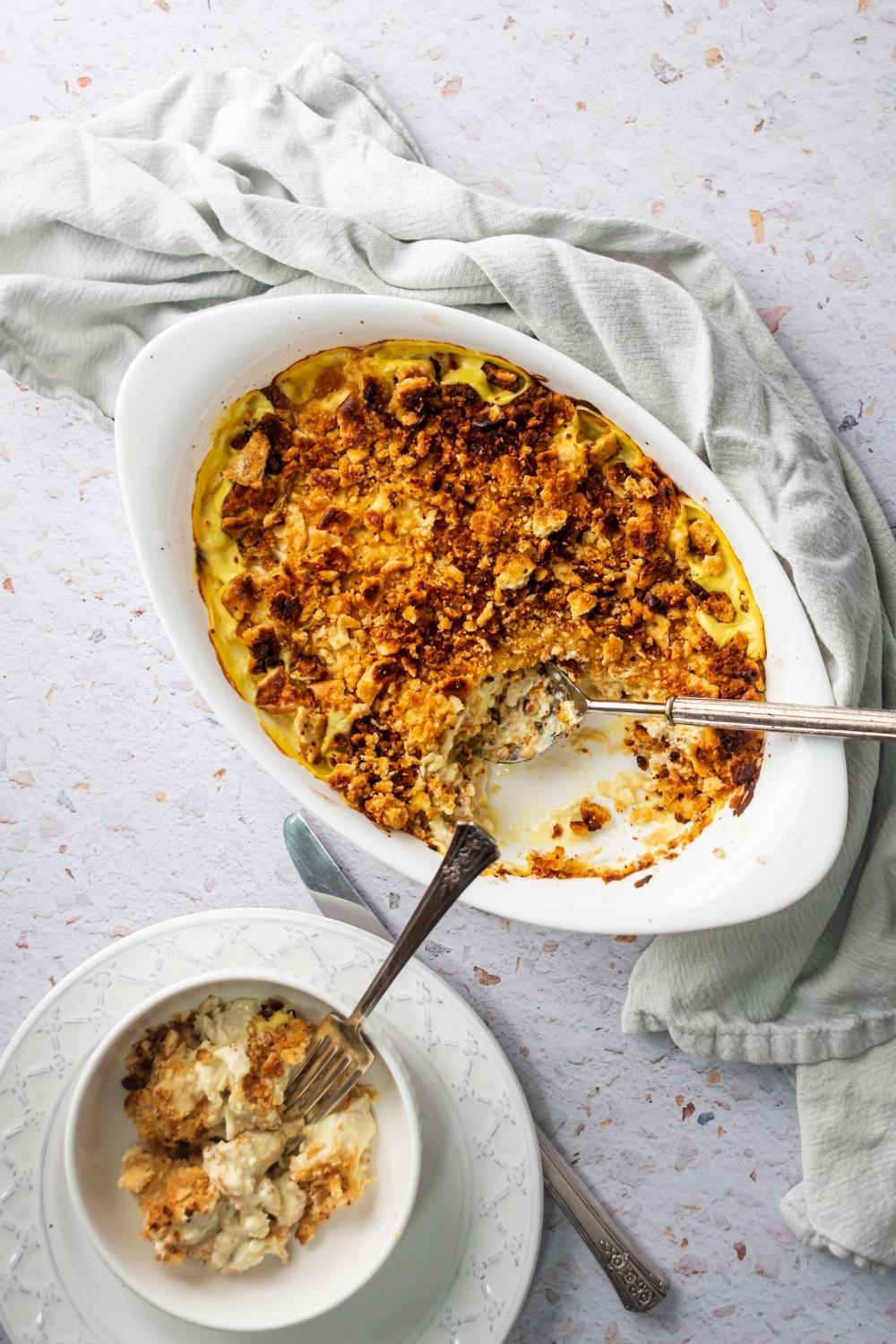 Million dollar chicken casserole in a white casserole dish. A sixth of the casserole is taken out of the front of it and it is in a white bowl that is on a white plate behind the casserole.