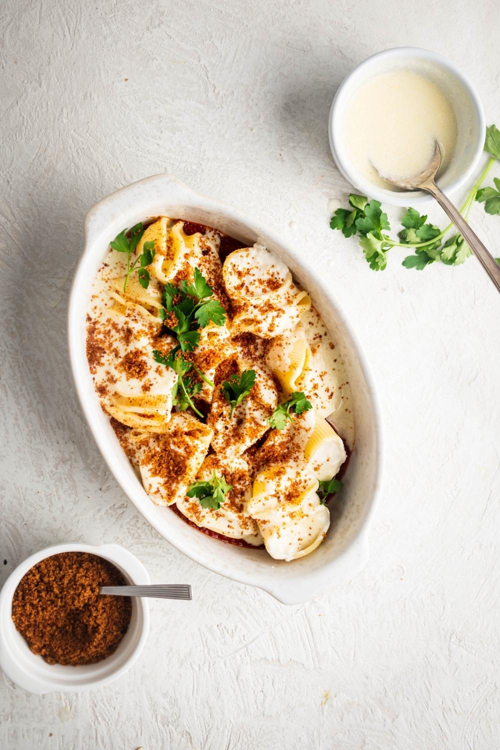 Giant cheese stuffed shells in a white casserole dish. In front of it is a bowl of toasted bread crumbs or behind it a bowl of Alfredo sauce..