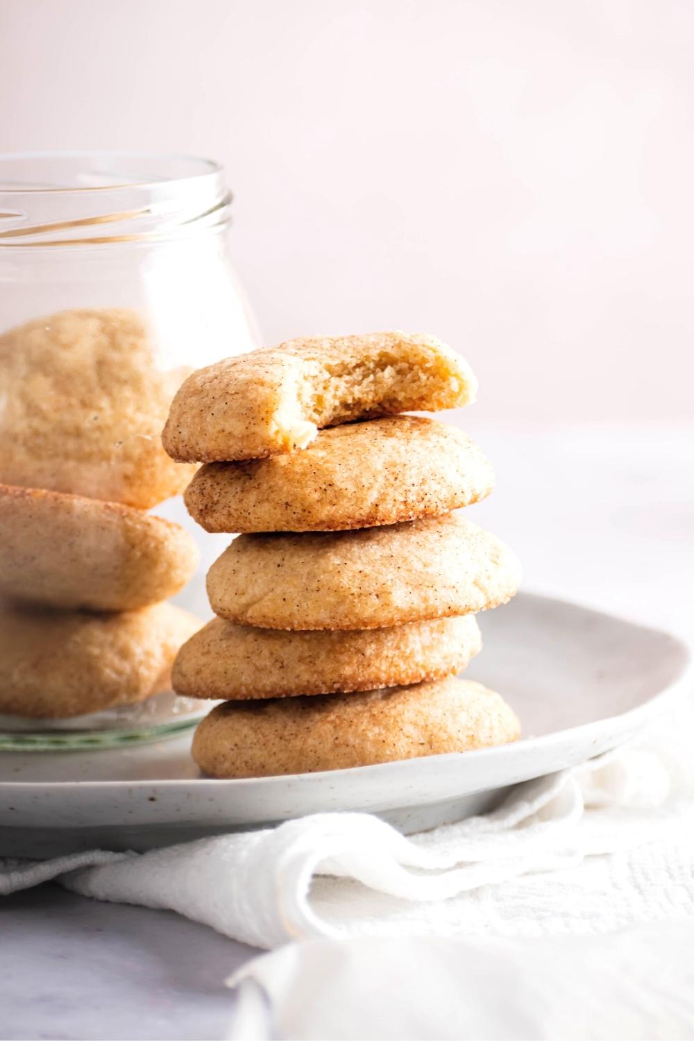 Five snickerdoodles without cream of tartar stacked on top of one another and a white plate. The top cookie has a bite out of the front of it in behind the staff is a glass jar filled with more cookies.