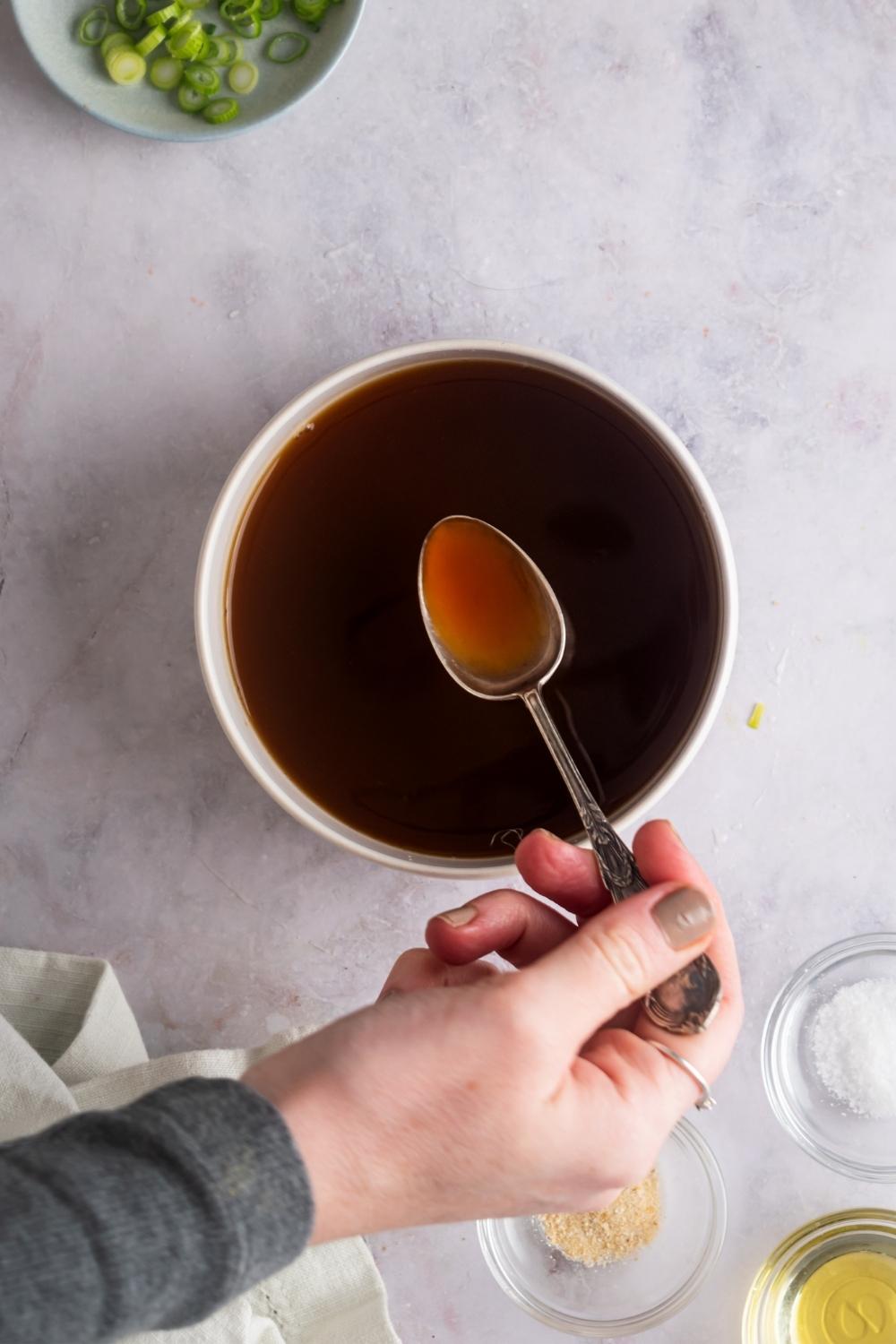 Hand holding a spoon that has a bourbon glaze on it being held above the bourbon glaze in a white bowl on a gray counter..