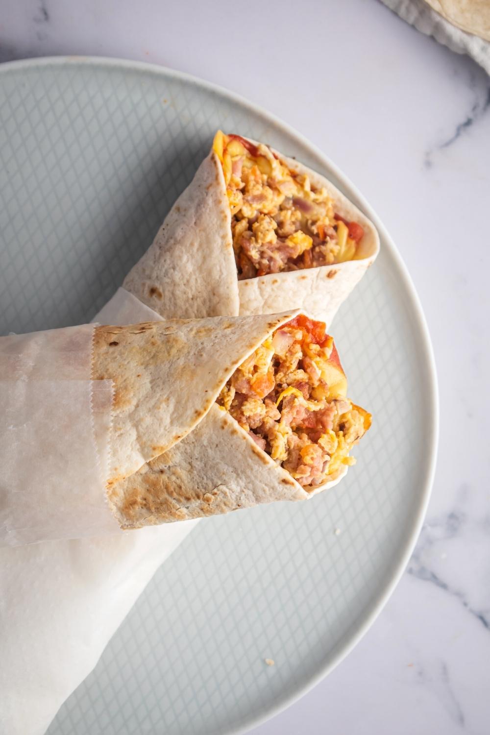 Half of a breakfast burrito wraps in parchment paper on top of another one of the breakfast burritos on part of a plate.