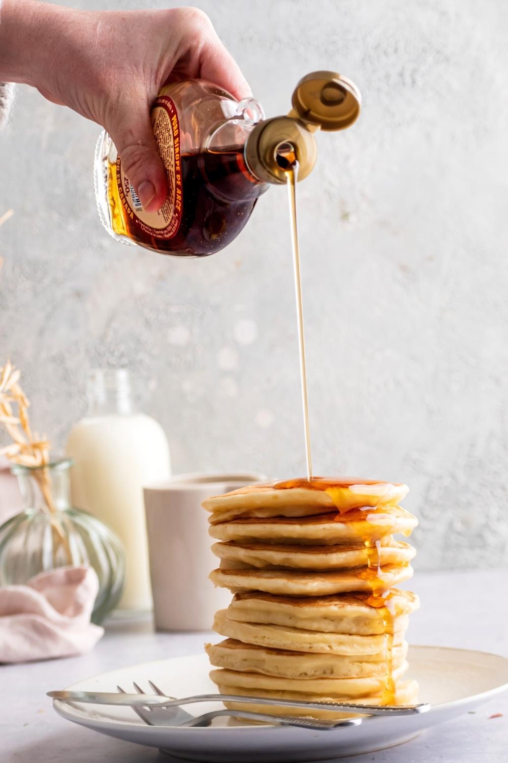 A hand holding a bottle of maple syrup pouring it on a stack of pancakes that are on a white plate with a fork and knife on it.