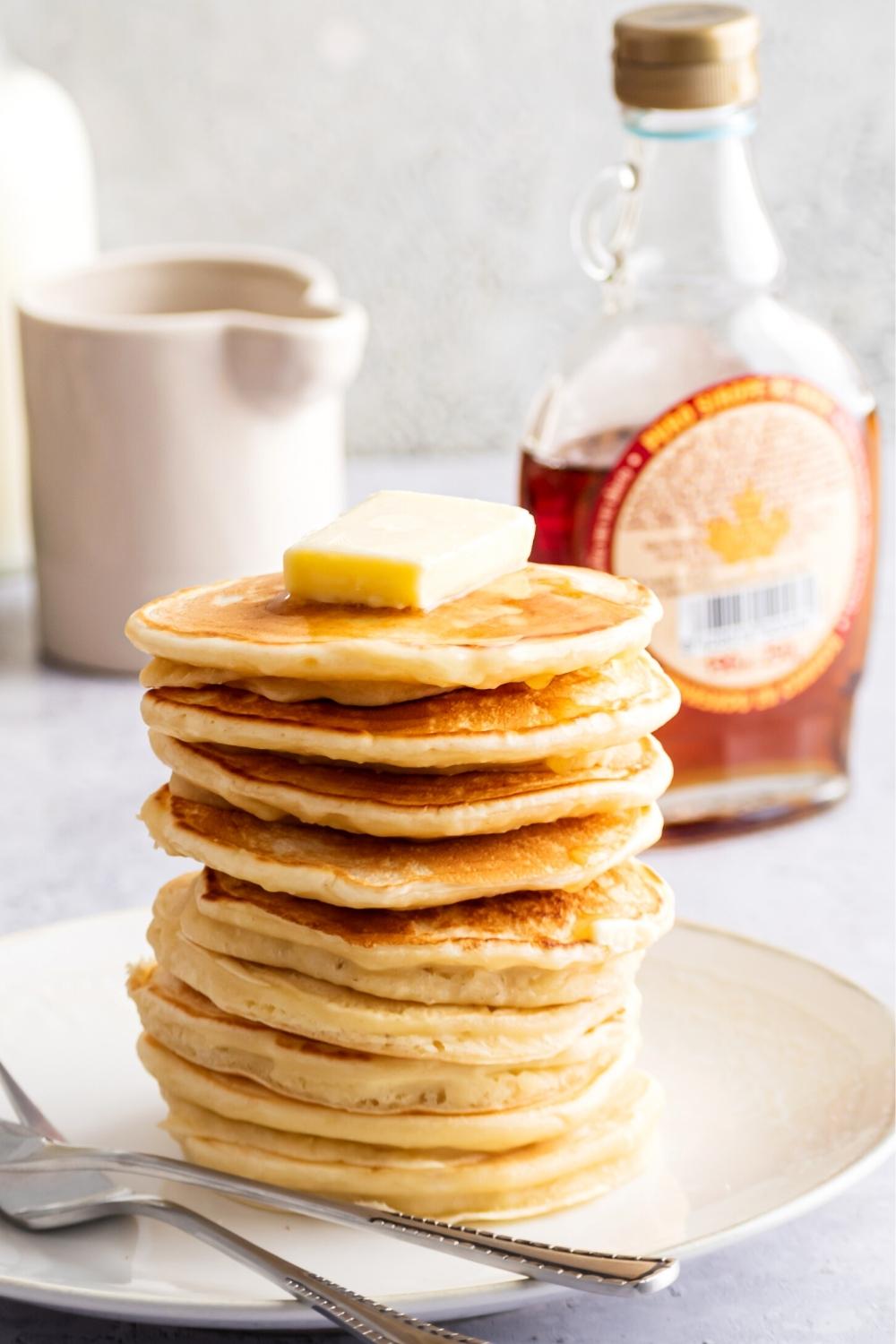 A stack of nine pancakes on a white plate. The top pancake has a square of butter on it. Behind the plate is a bottle of maple syrup.