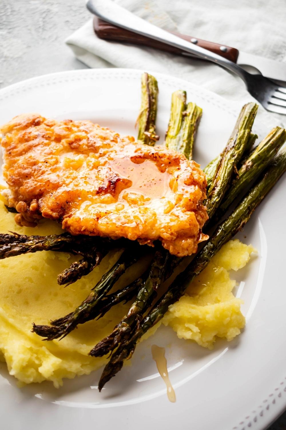 Truffle honey on top of a piece of breaded chicken that is an asparagus spears on mashed potatoes on a white plate.