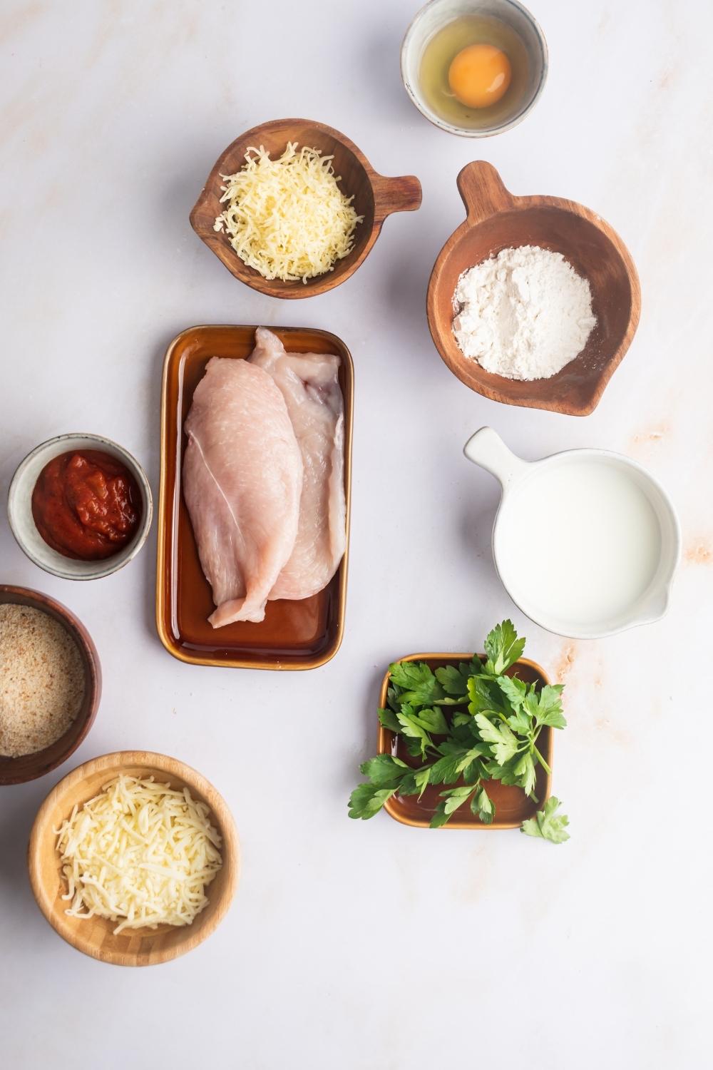 A bowl of shredded mozzarella cheese, part of a bowl of breadcrumbs, both marinara sauce in it, two slices of chicken breast on a plate, bowl of mozzarella cheese, bowl with an egg in it, bowl flour in it, and a bowl with milk in it all on a white counter.