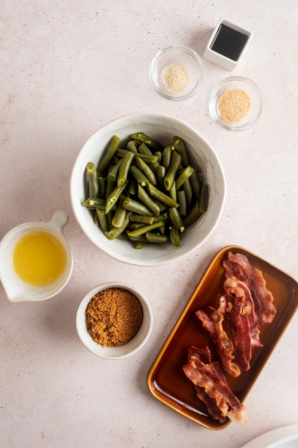 A bowl of green beans, a plate of bacon, bowl brown sugar, both melted butter, bowl garlic powder, bowl of onion powder, and evolve tamari sauce on the white counter.