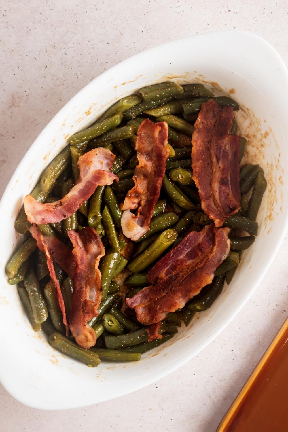 Six slices of bacon on top of green beans and a white casserole dish.