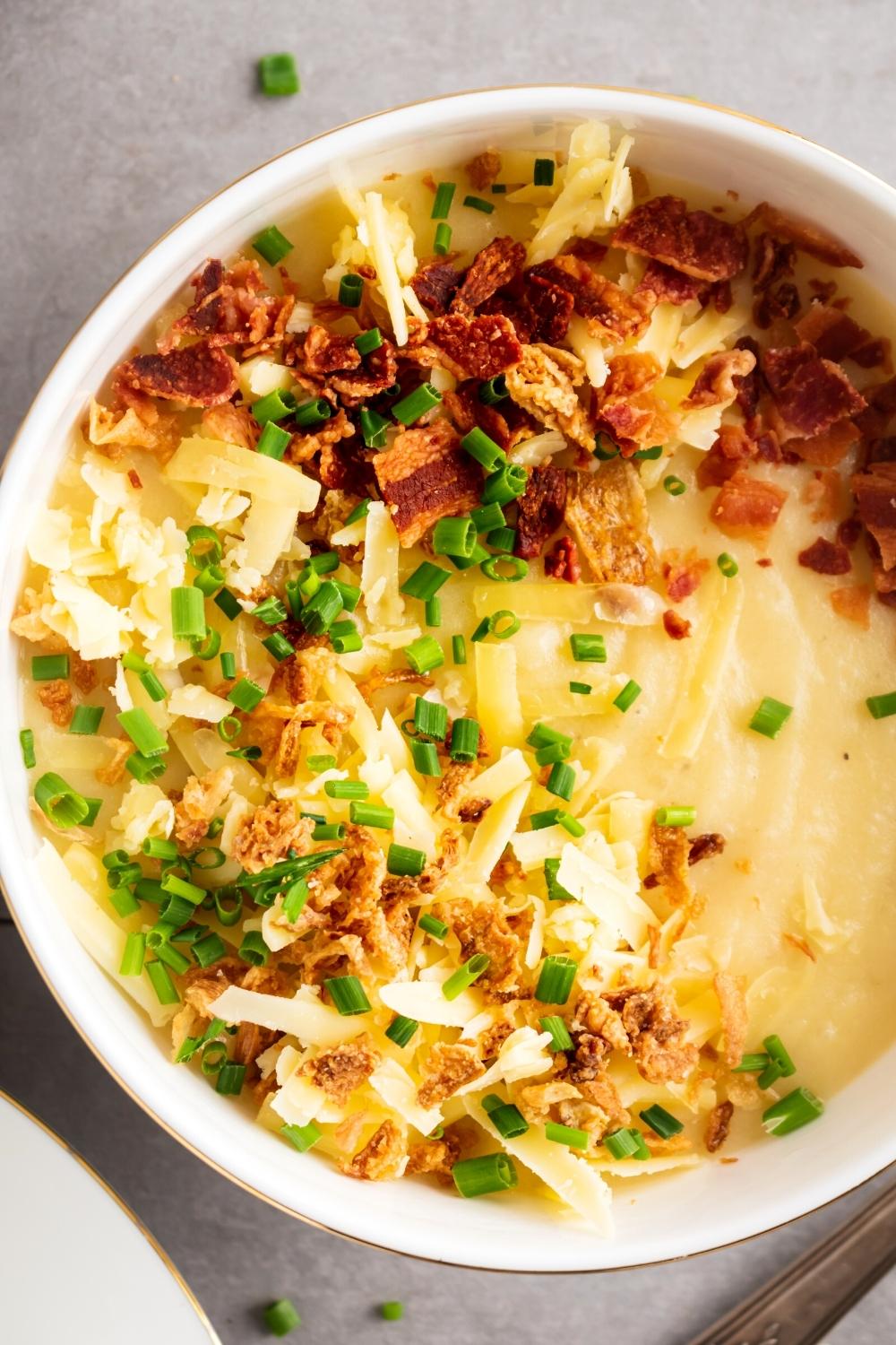 Shredded cheese, bacon bits, and crispy onions on top of potato soup in a white bowl.