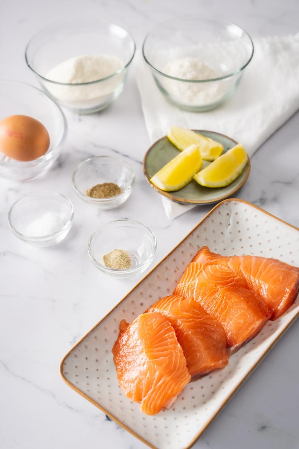 Four salmon fillets on a rectangular plate, a bowl of salt, a bowl of pepper, and a bowl of seasoning are behind it.