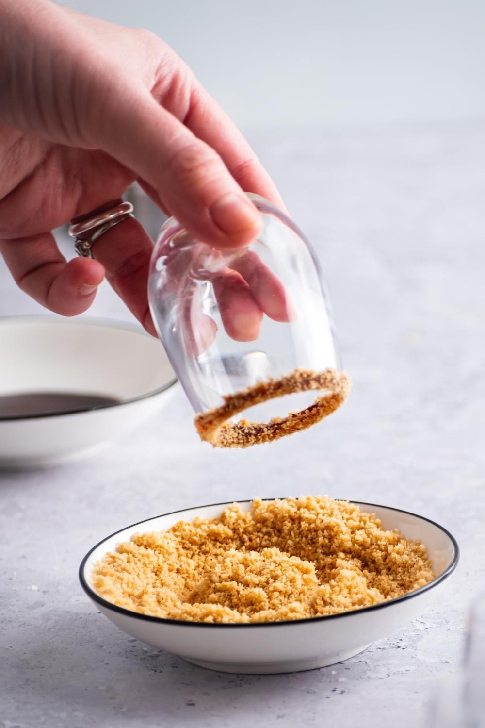 A hand holding a shot glass upside down with crushed up graham crackers on the rim of it. The shot glass is being held above a bowl that is filled with the crushed up graham crackers.