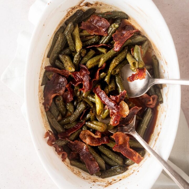 Krispy pieces of bacon on top of green beans on two spoons in a white casserole dish.
