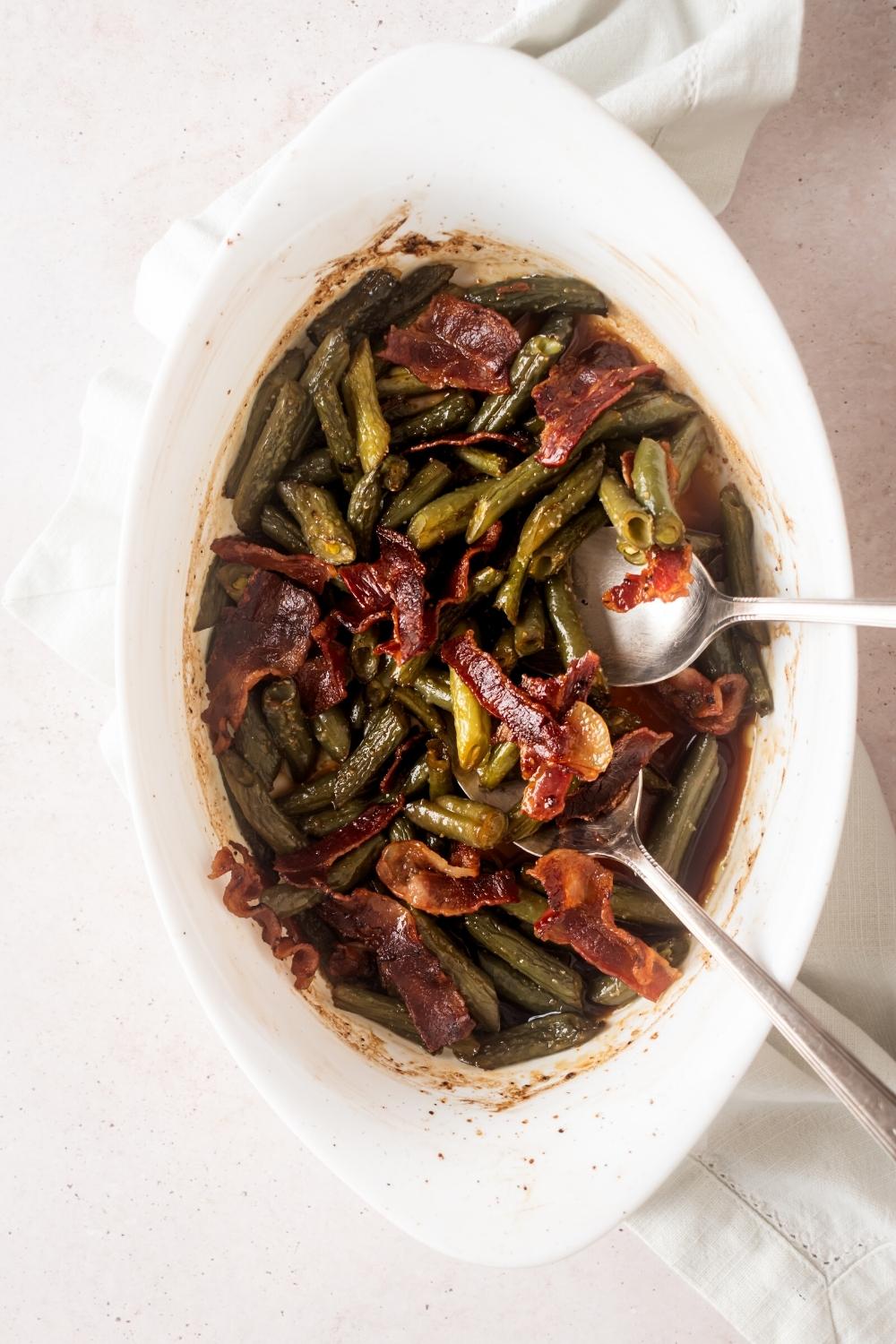 Krispy pieces of bacon on top of green beans on two spoons in a white casserole dish.