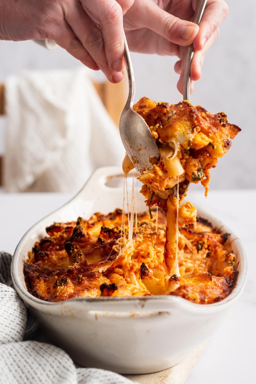 Two hands call Dean utensils with four cheese pasta between them being pulled from a white casserole dish that is filled with the four cheese pasta.