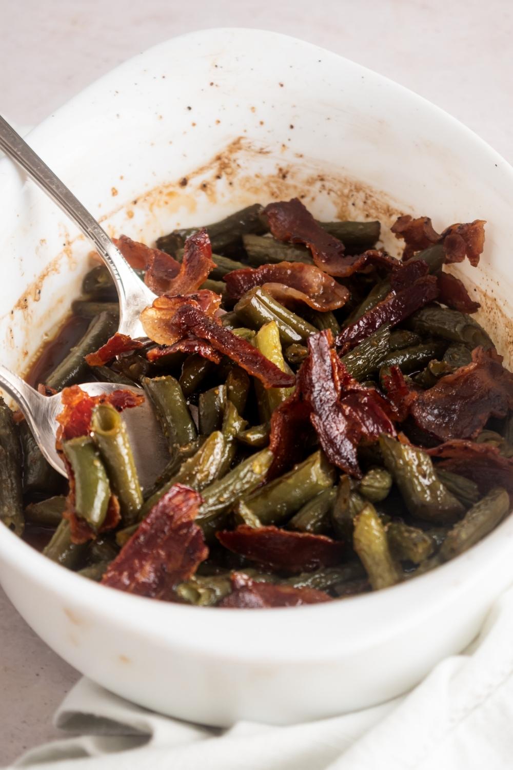 Krispy pieces of bacon on top of green beans in a white casserole dish.