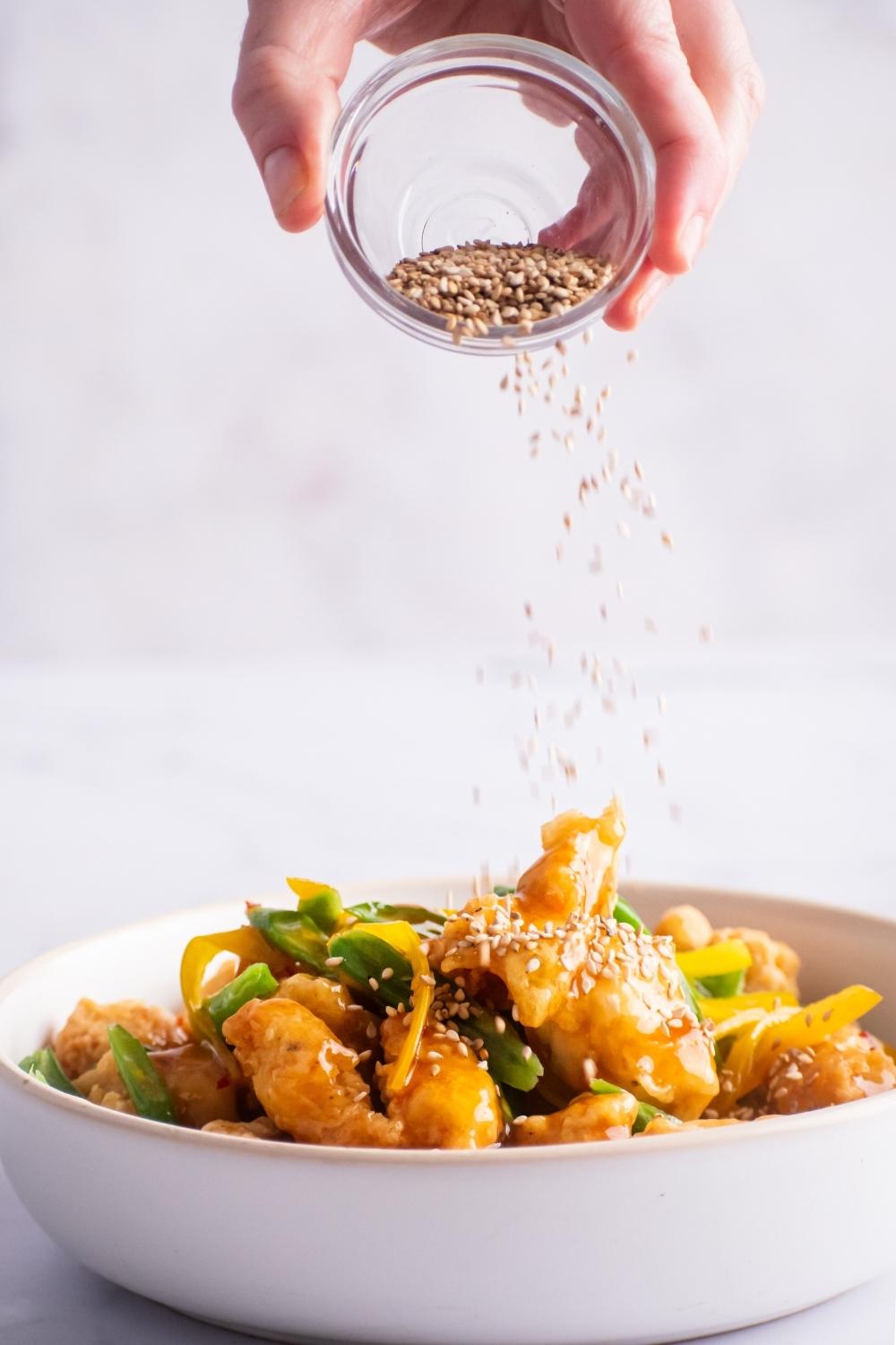 Hand sprinkling sesame seeds on top of honey sesame chicken, green beans, and yellow bell peppers in a white bowl.