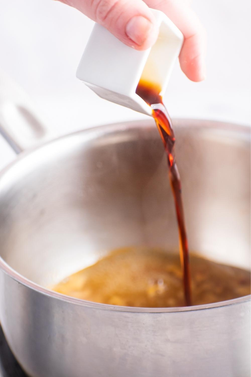 Hand pouring a square dish of soy sauce in a pot.