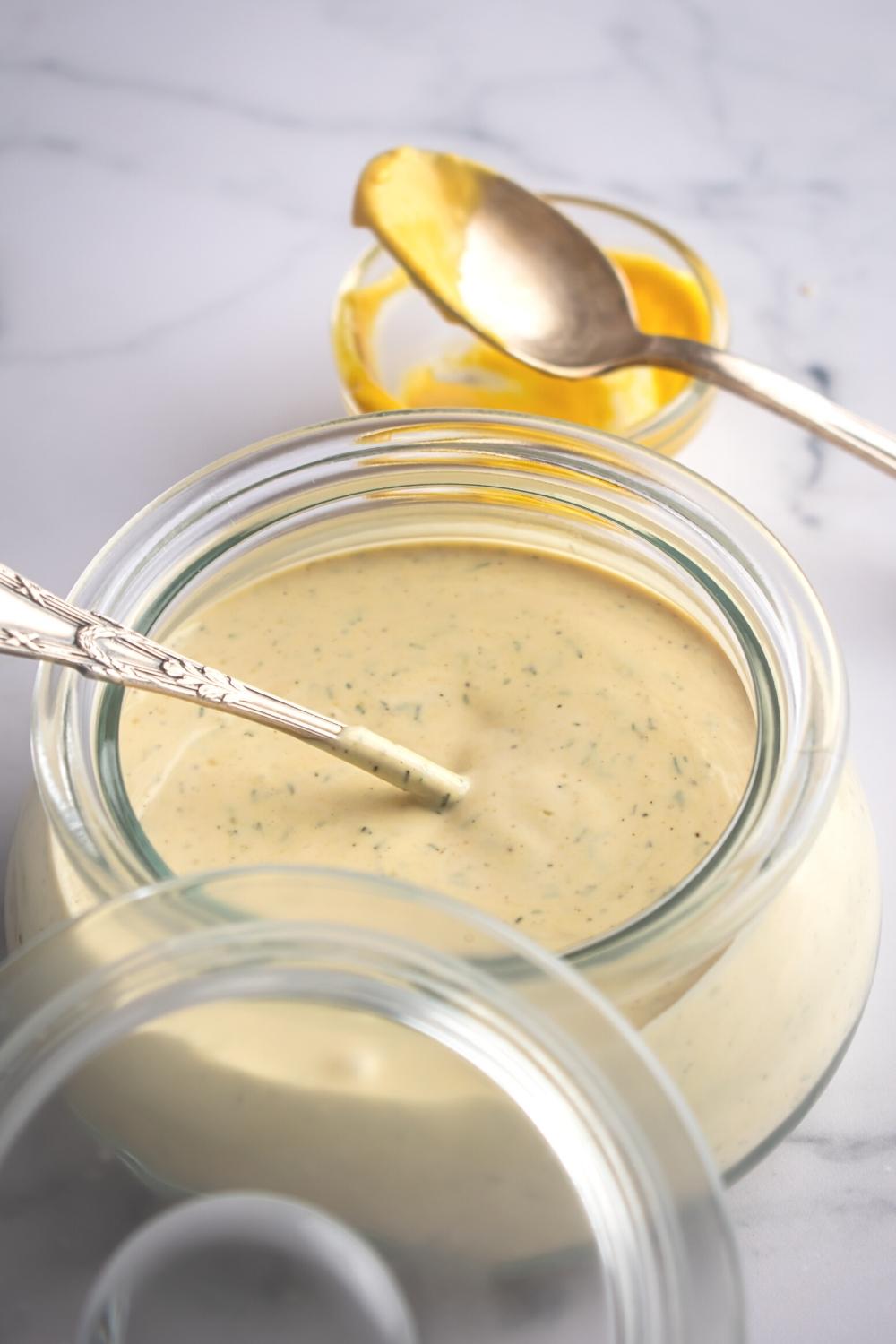 McDonald's breakfast sauce in a glass jar with a spoon submerged in the sauce. Behind it is a glass cup filled with mustard with a spoon on top.