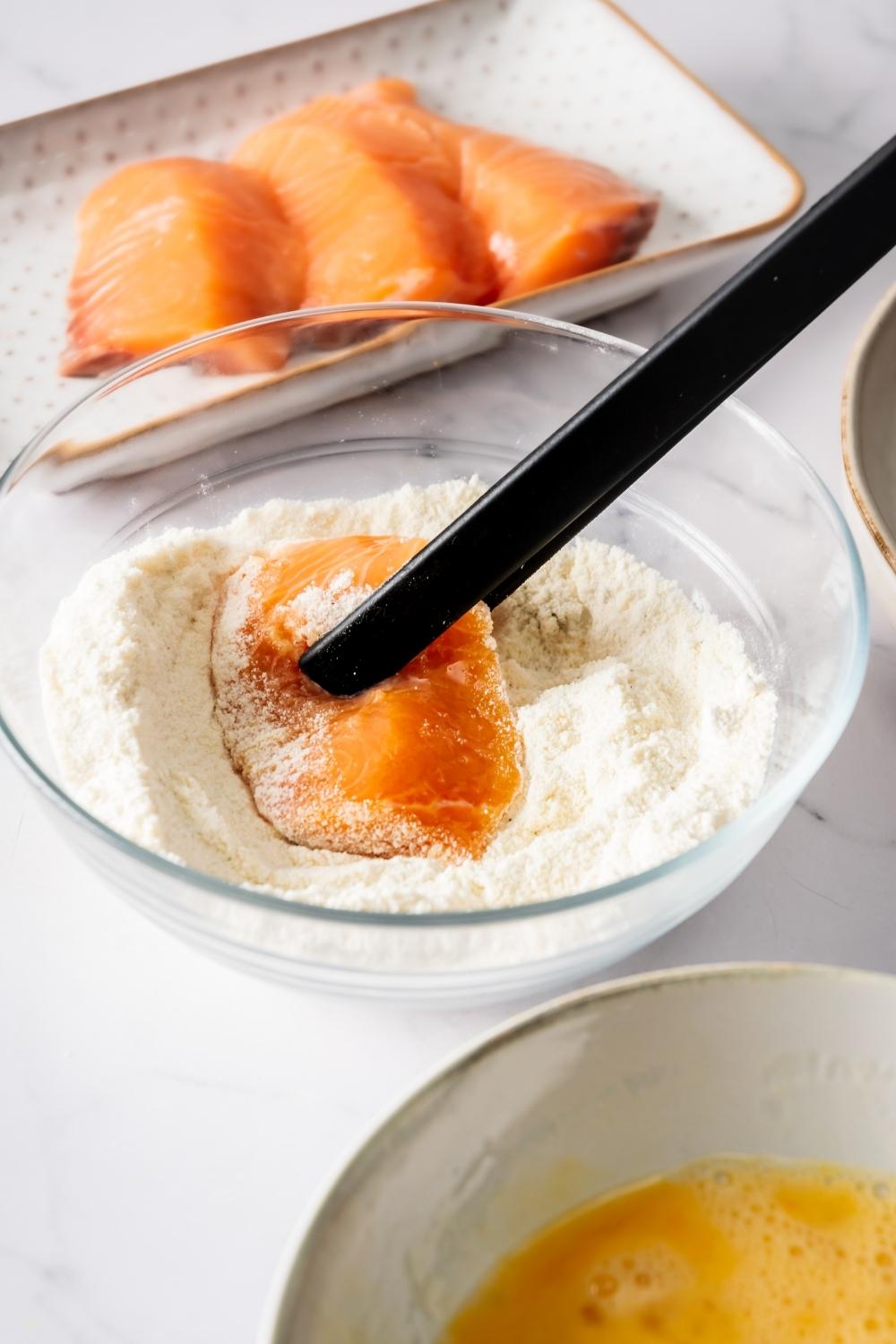 A piece of salmon in a bowl of flour with tongs clasping the fillet. Behind it is a plate with three salmon fillets on it.