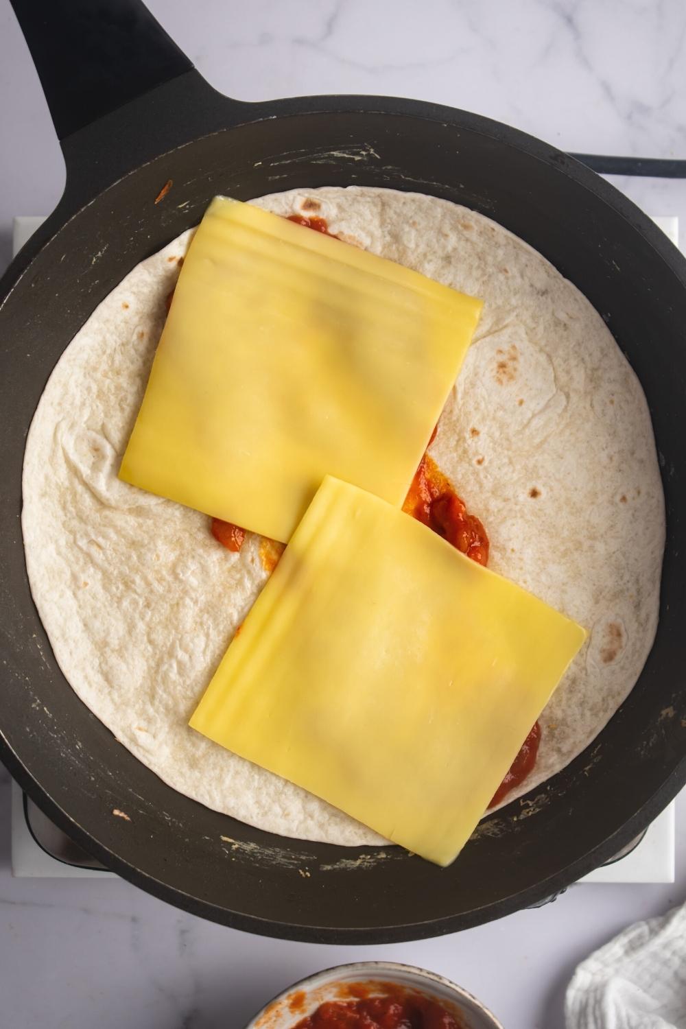 Slices of American cheese on top of salsa that is in the middle of a flour tortilla in a skillet.