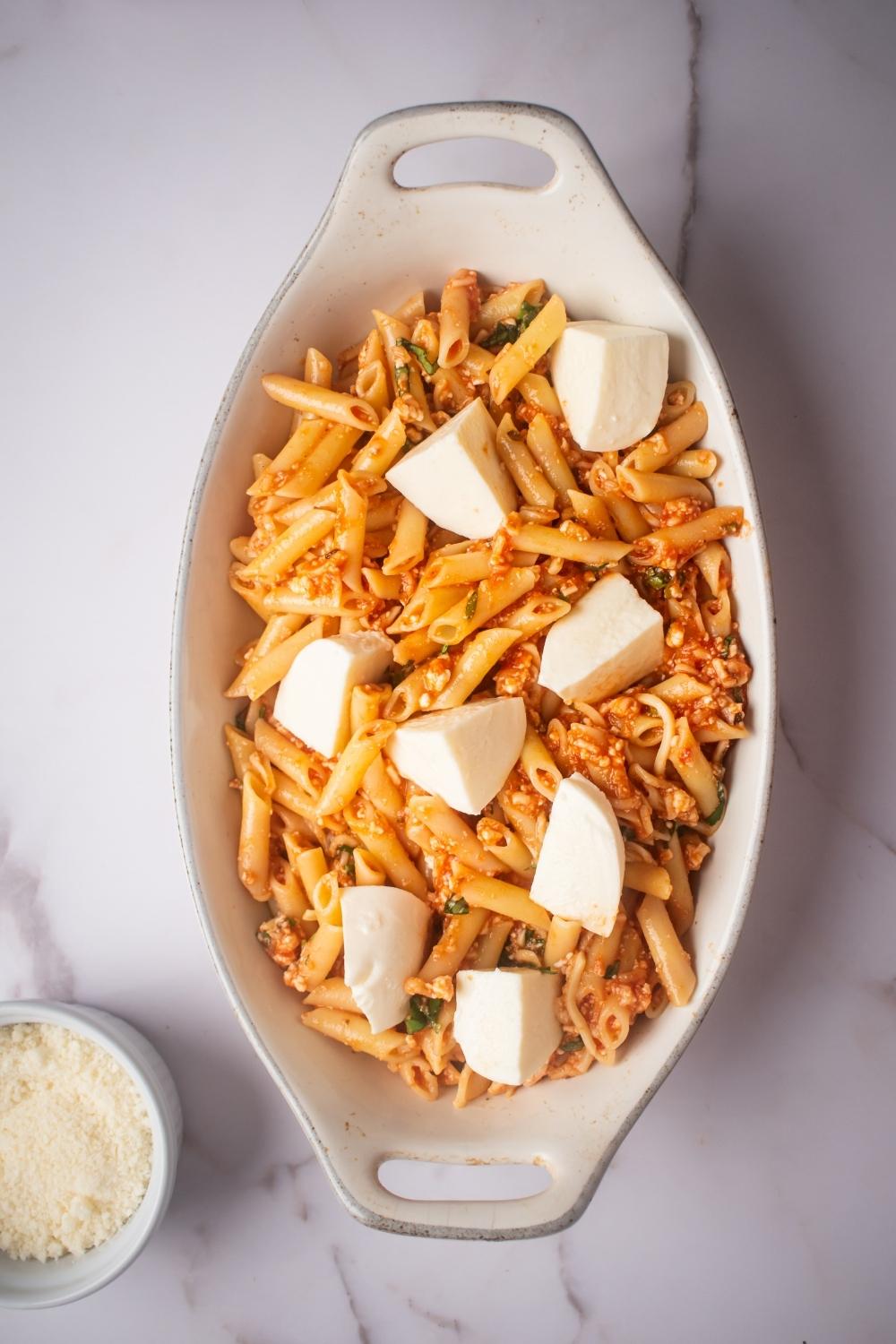 Fresh mozzarella cubes on top of penne pasta in a white baking dish.