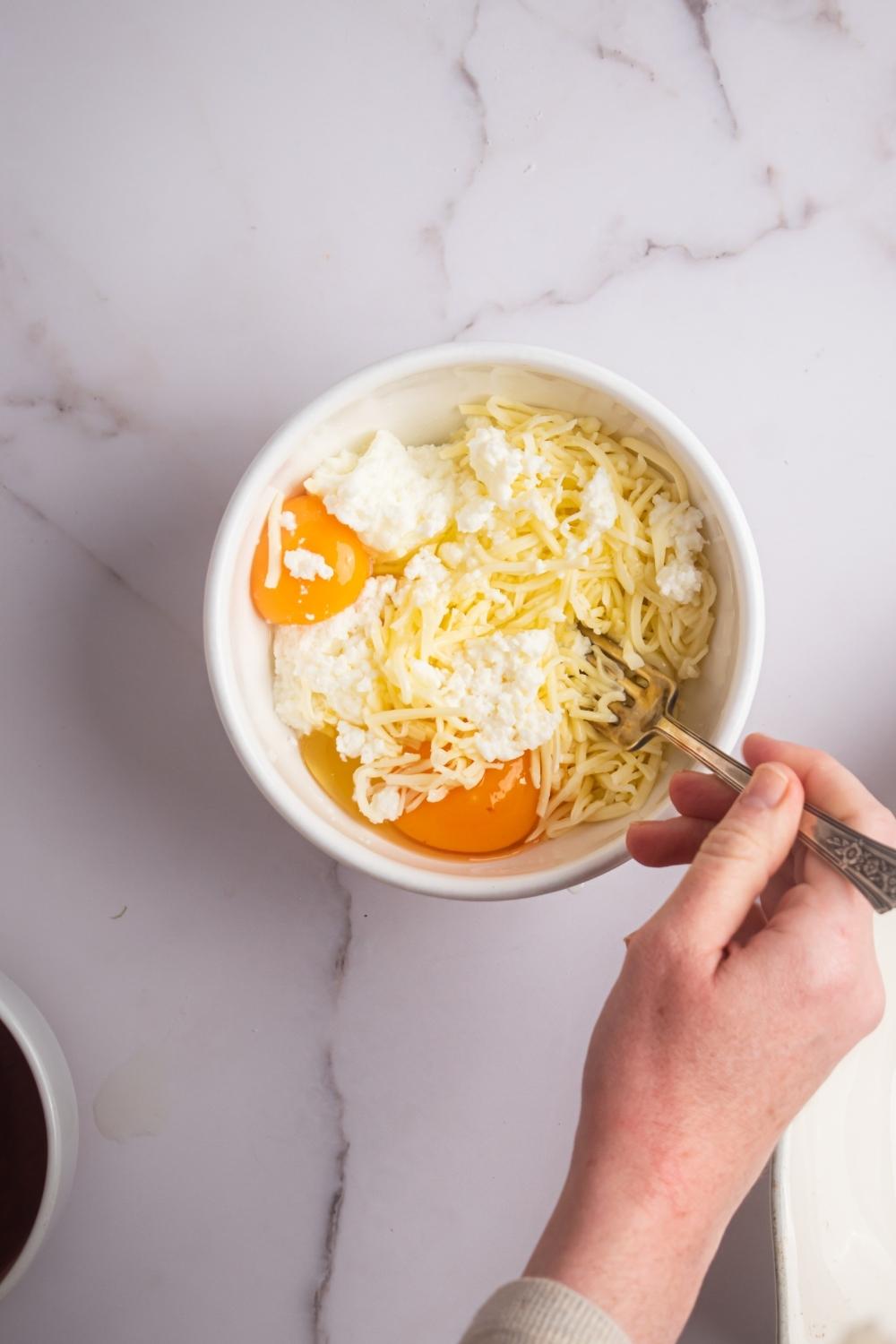 A hand holding a fork that is submerged in a white bowl filled with two eggs, ricotta cheese, and shredded Parmesan cheese.