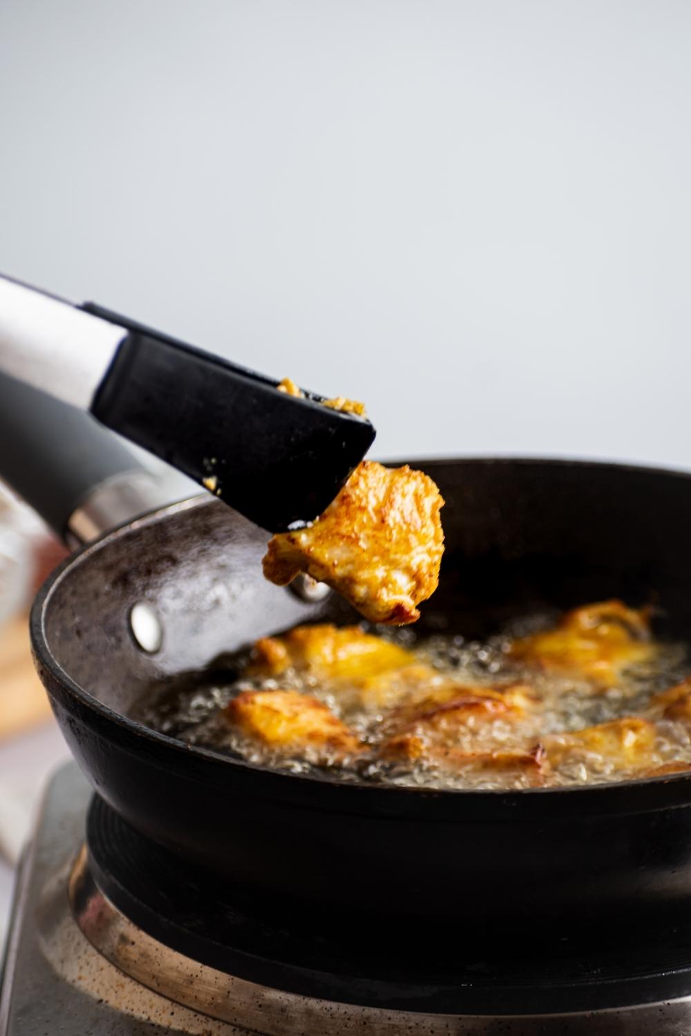 Tongs with a piece of piece of chicken between them over a pan that has chicken frying in it.
