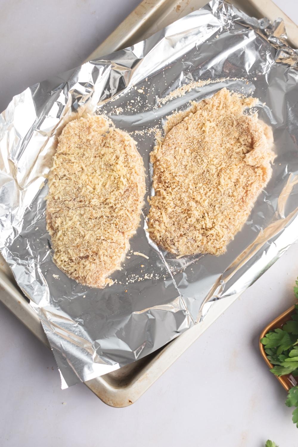 Two pieces of raw breaded chicken on a sheet of tinfoil on a baking tray.
