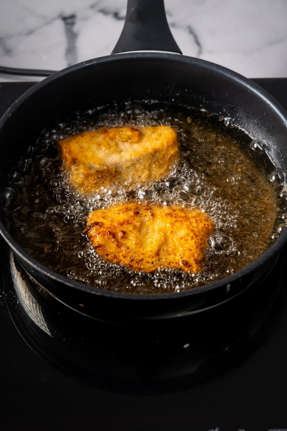 Two pieces of fried salmon in a pan that is filled with oil.