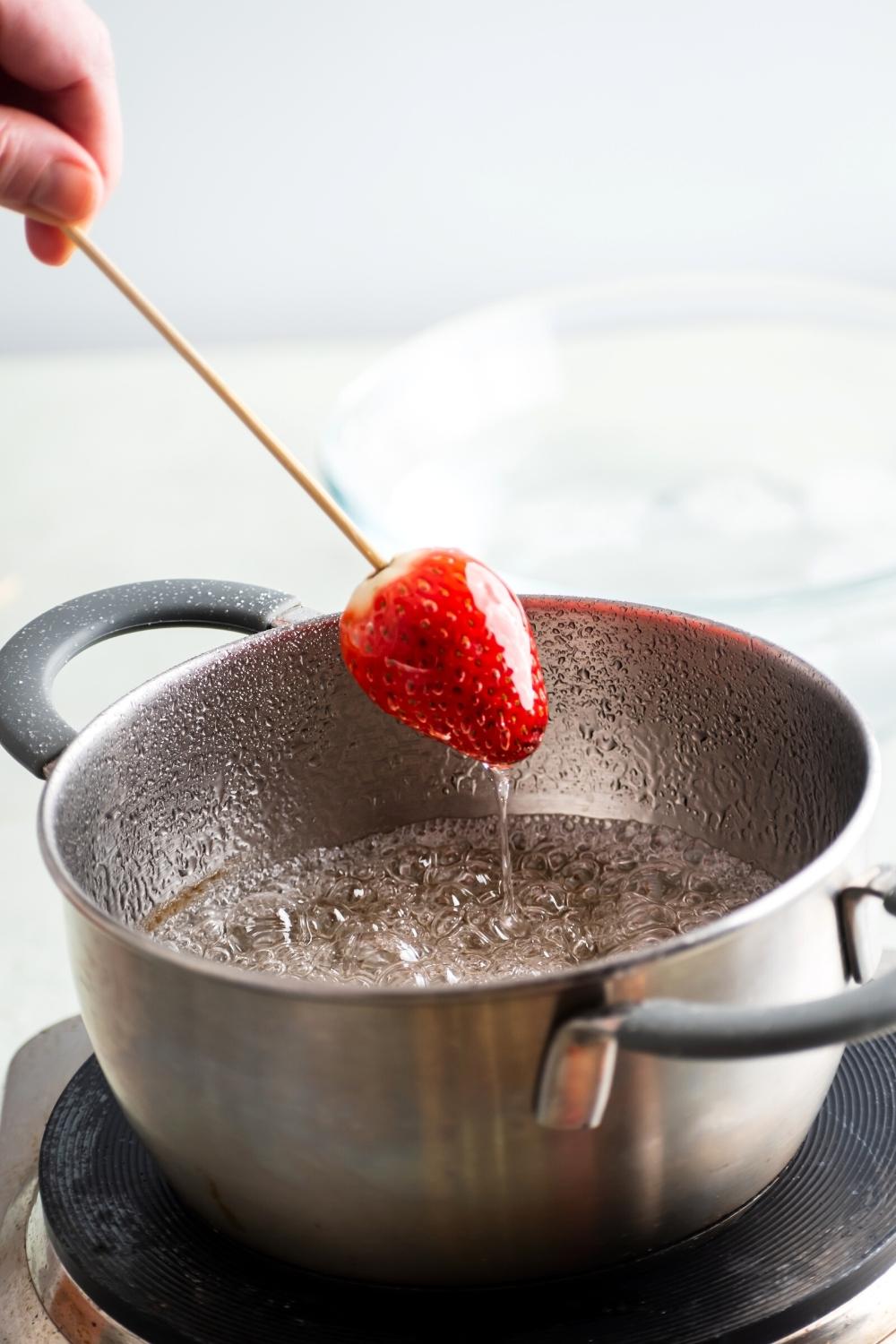 A strawberry on a wooden skewer being held over apart filled with water and sugar.