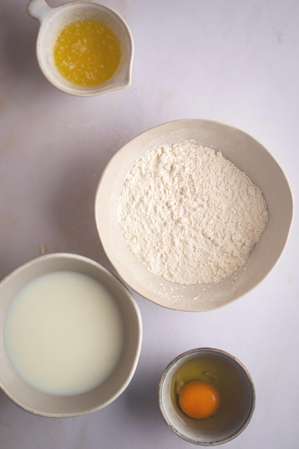 Flower and white bowl, milk and white bowl, an egg in a bowl, and oil in a ball on a white counter