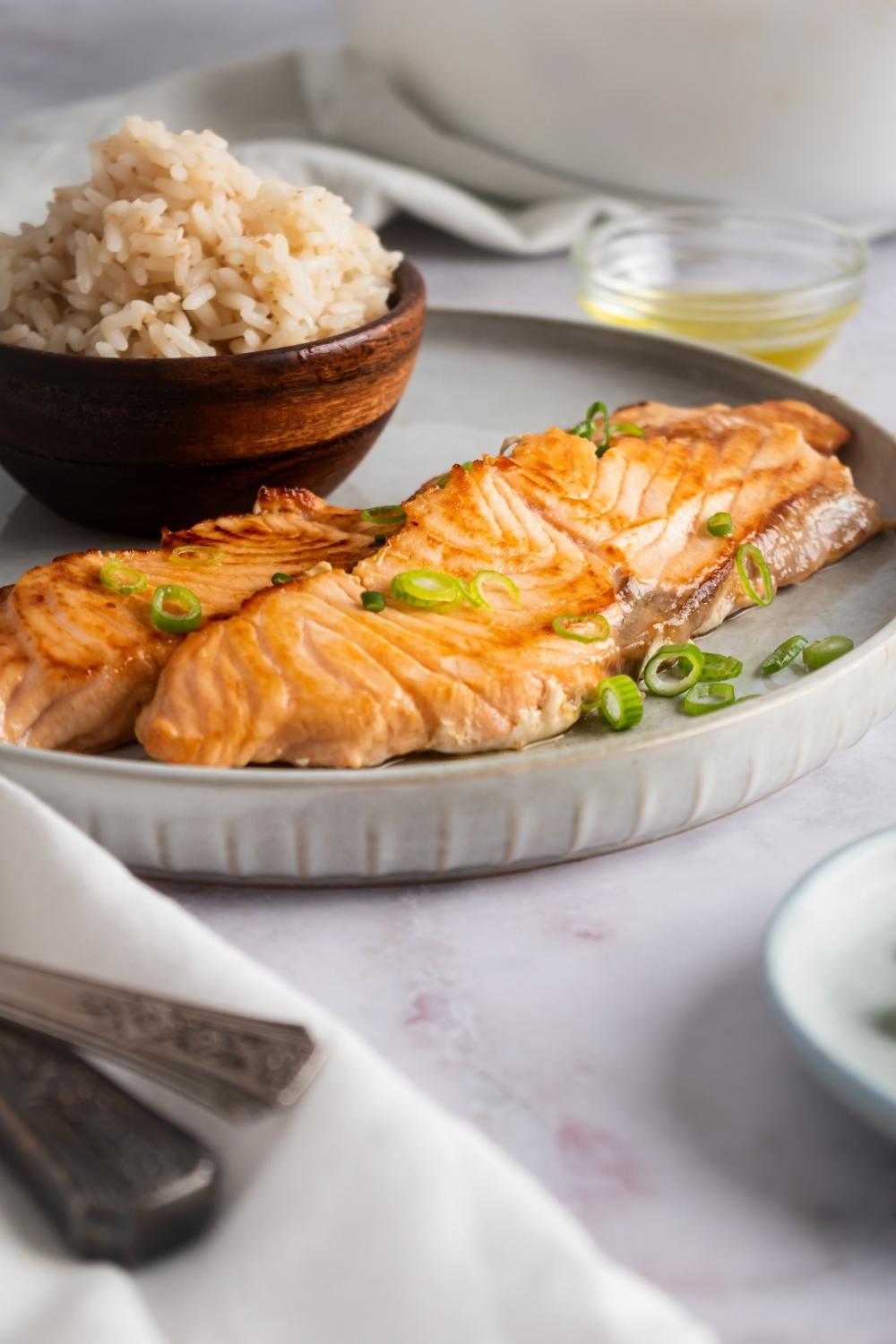 Two fillets of longhorn salmon on a gray plate that has a wooden bowl filled with rice on it.