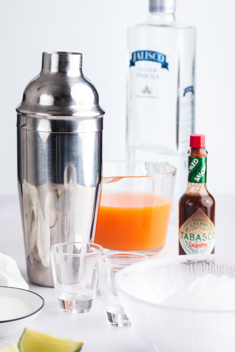 A cocktail mixer, two shot glasses, a bottle of tobasco, a jug of fruit juice, and a bottle of vodka.