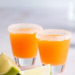 Two mexican candy shots with sugar on the rims of the shot glasses. In front of the glasses is two lime wedges.