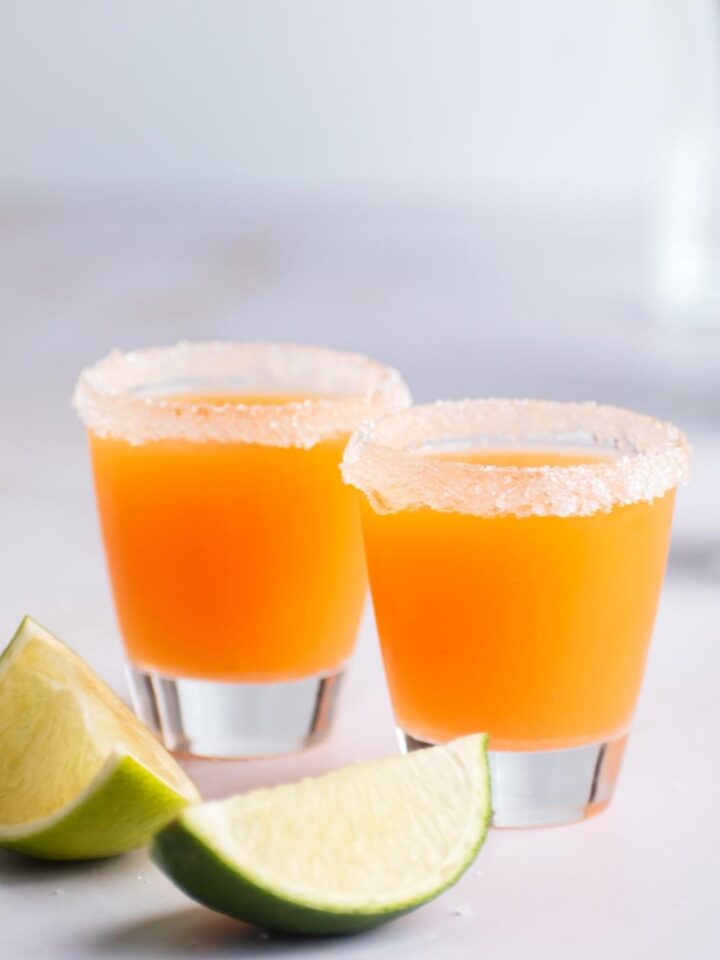 Two mexican candy shots with sugar on the rims of the shot glasses. In front of the glasses is two lime wedges.