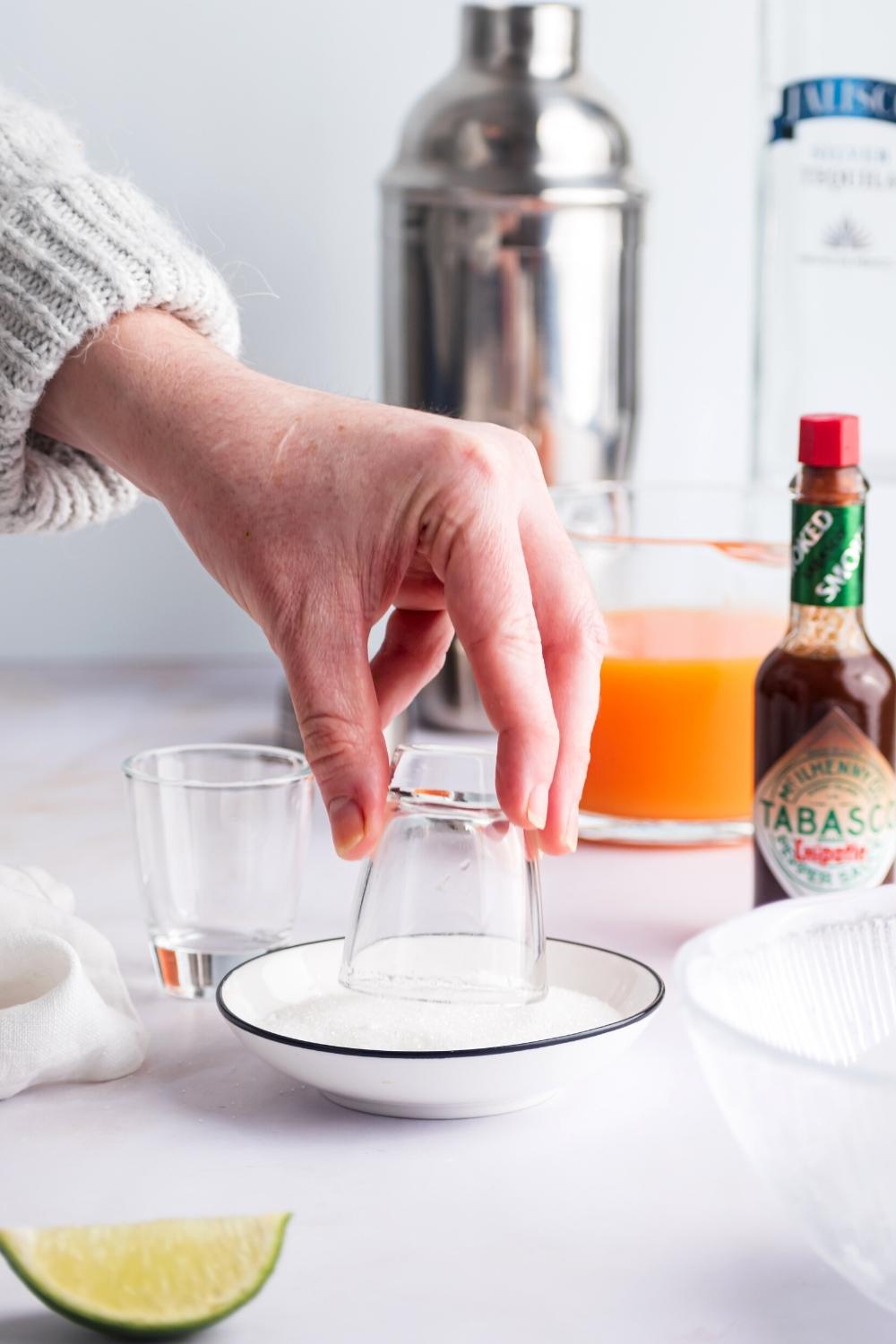 A hand dipping a shot glass into a bowl of sugar. Behind it is a jug of fruit juice, tobasco, and a cocktail shaker.