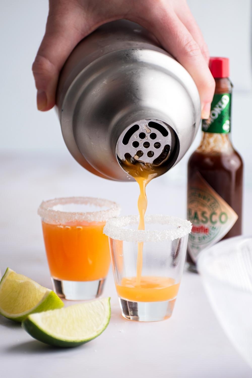 A hand pouring a mexican candy shot into a shot glass with sugar on the rim. Behind it is another mexican lollipop shot and a bottle of tobasco.