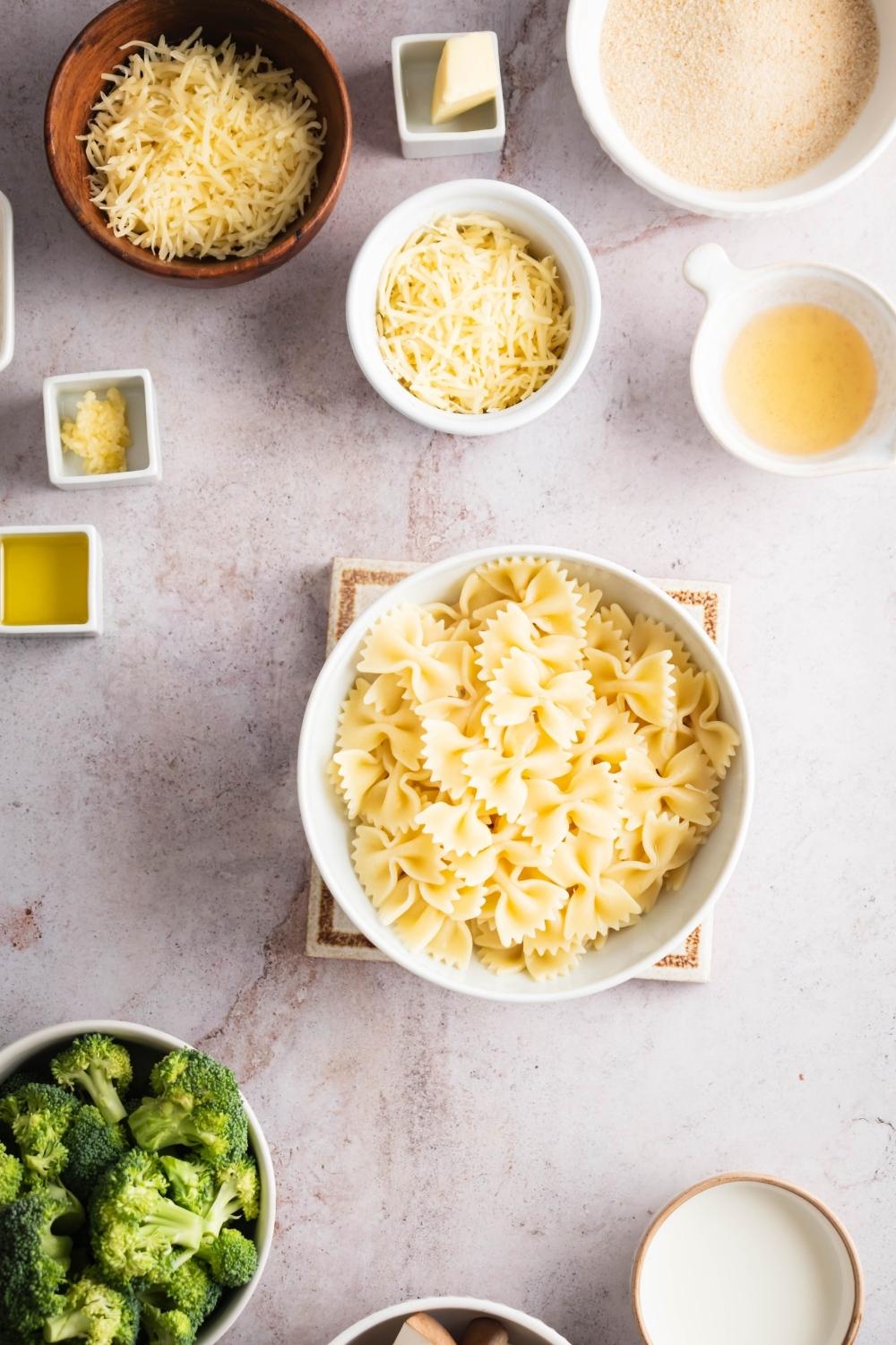 Bow tie pasta in a bowl surrounded by a bowl of parmesan cheese, a bowl of asiago cheese, a bowl of broccoli, and a bowl of bread crumbs.