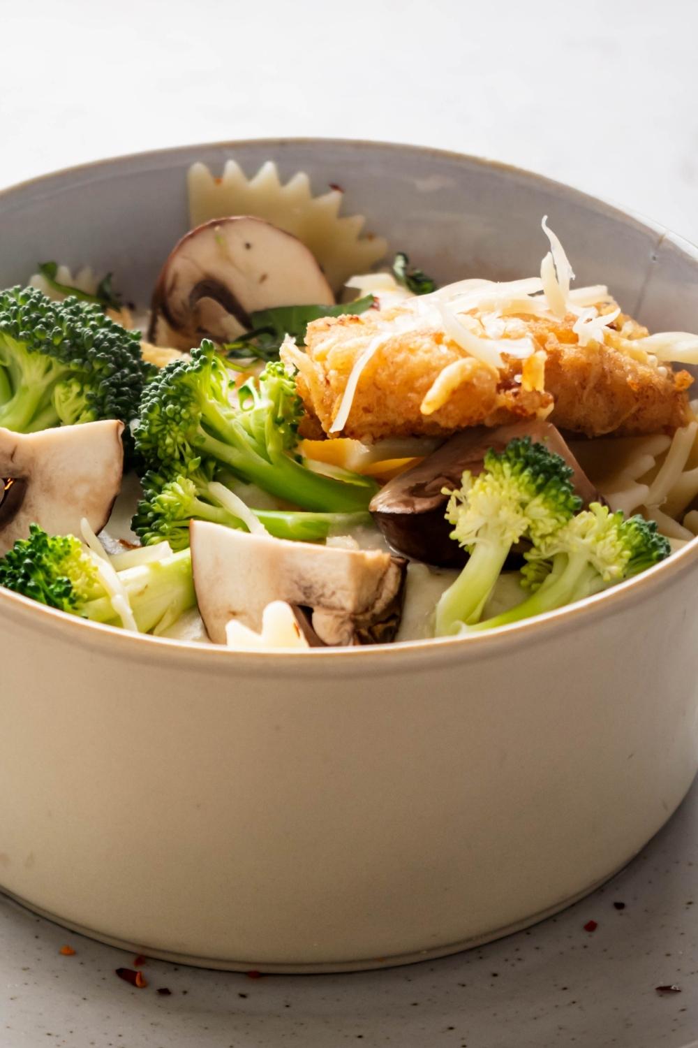 A bowl filled with broccoli, a piece of crispy chicken fritta, mushrooms, and bow tie pasta.