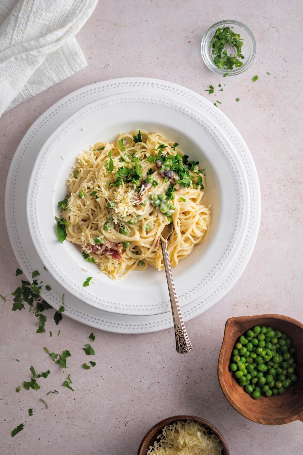 Pasta carbonara and white bowl on top of white plate on a white counter. In front of the bowl is a bowl of peas