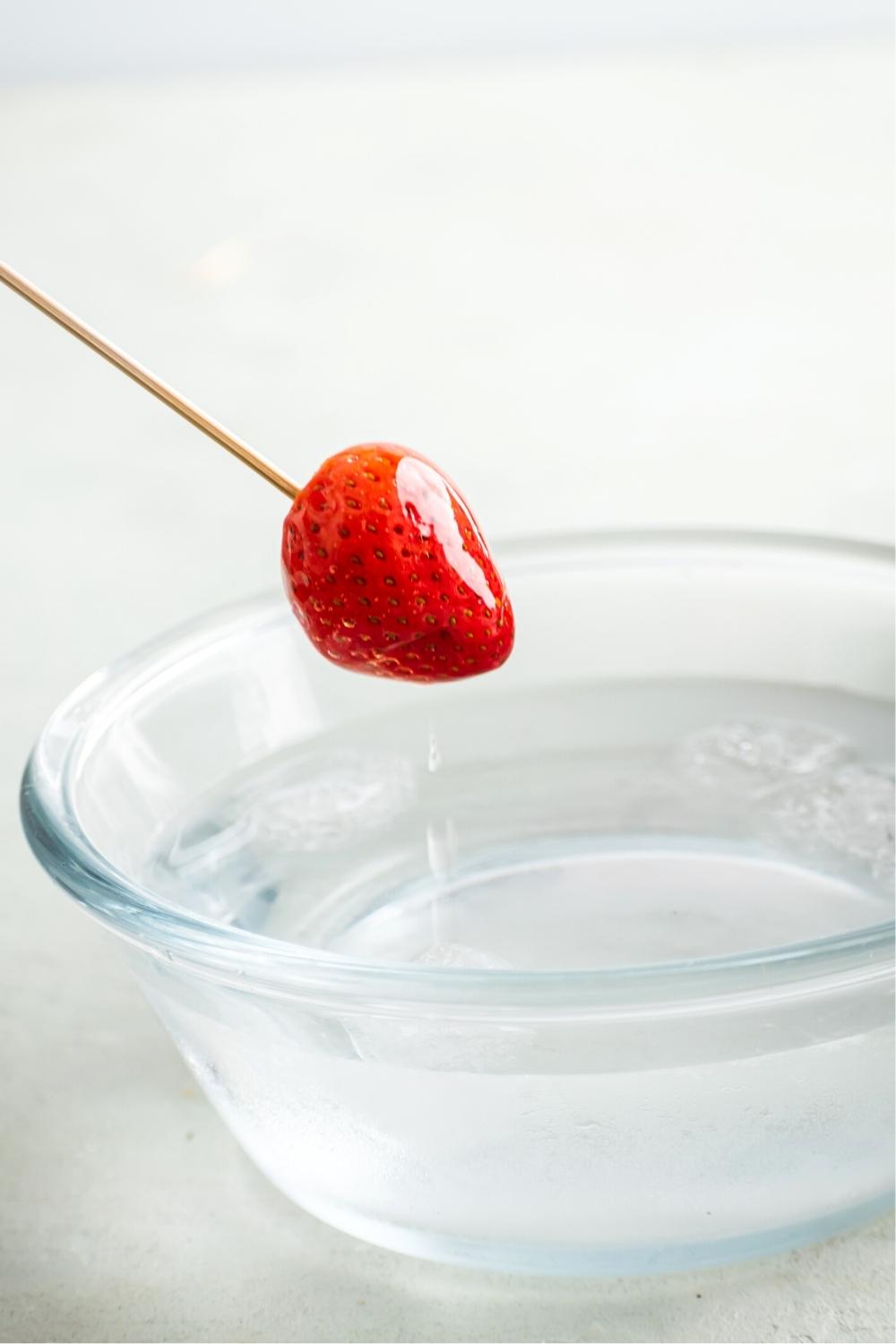 A strawberry on a wooden stick dripping water into a glass bowl filled with ice water.