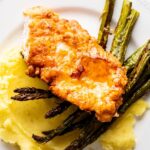 Shuffle honey on top of chicken that is an asparagus spears on top of mashed potatoes on a white plate.