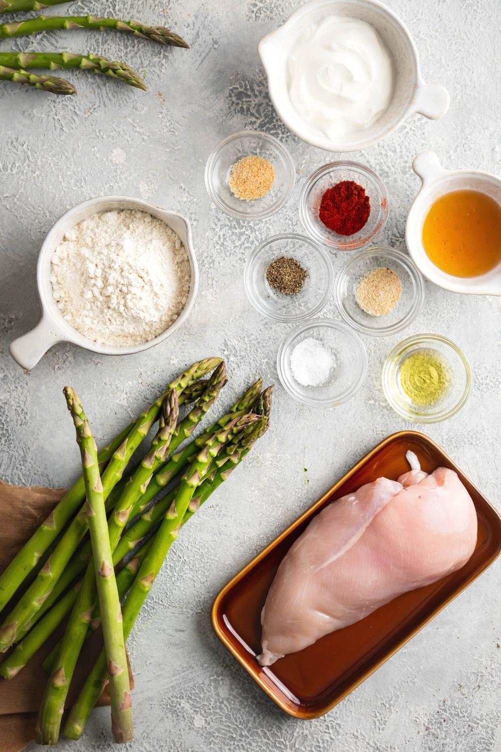 A chicken breast, a few spears of asparagus, a cup of garlic powder, cup of oil, a cup of paprika, cup of onion powder, Balflour, ball buttermilk, and a bowl truffle honey on a gray counter.