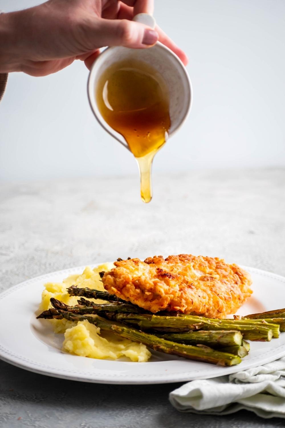 Hand pouring shuffle honey on top of a piece of breaded chicken that is on asparagus and mashed potatoes on a white plate.