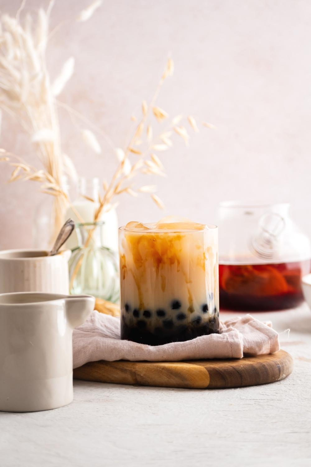 A cup of brown sugar milk tea on a napkin on a wood cutting board on a white counter. Behind it is a pitcher filled with black tea.