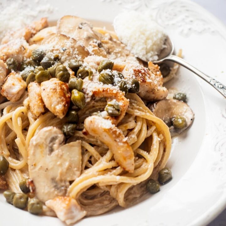 An overhead view of a white dish with cheesecake factory chicken piccata. A spoon with a heaping scoop of grated parmesan cheese.